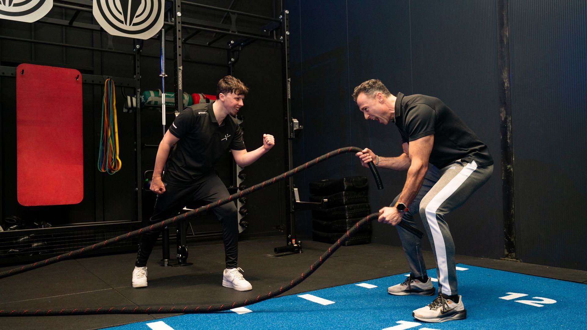 Mensen oefenen met gewichten in een sportschool; vrouw in een roze tanktop lacht terwijl ze een kettlebell tilt.