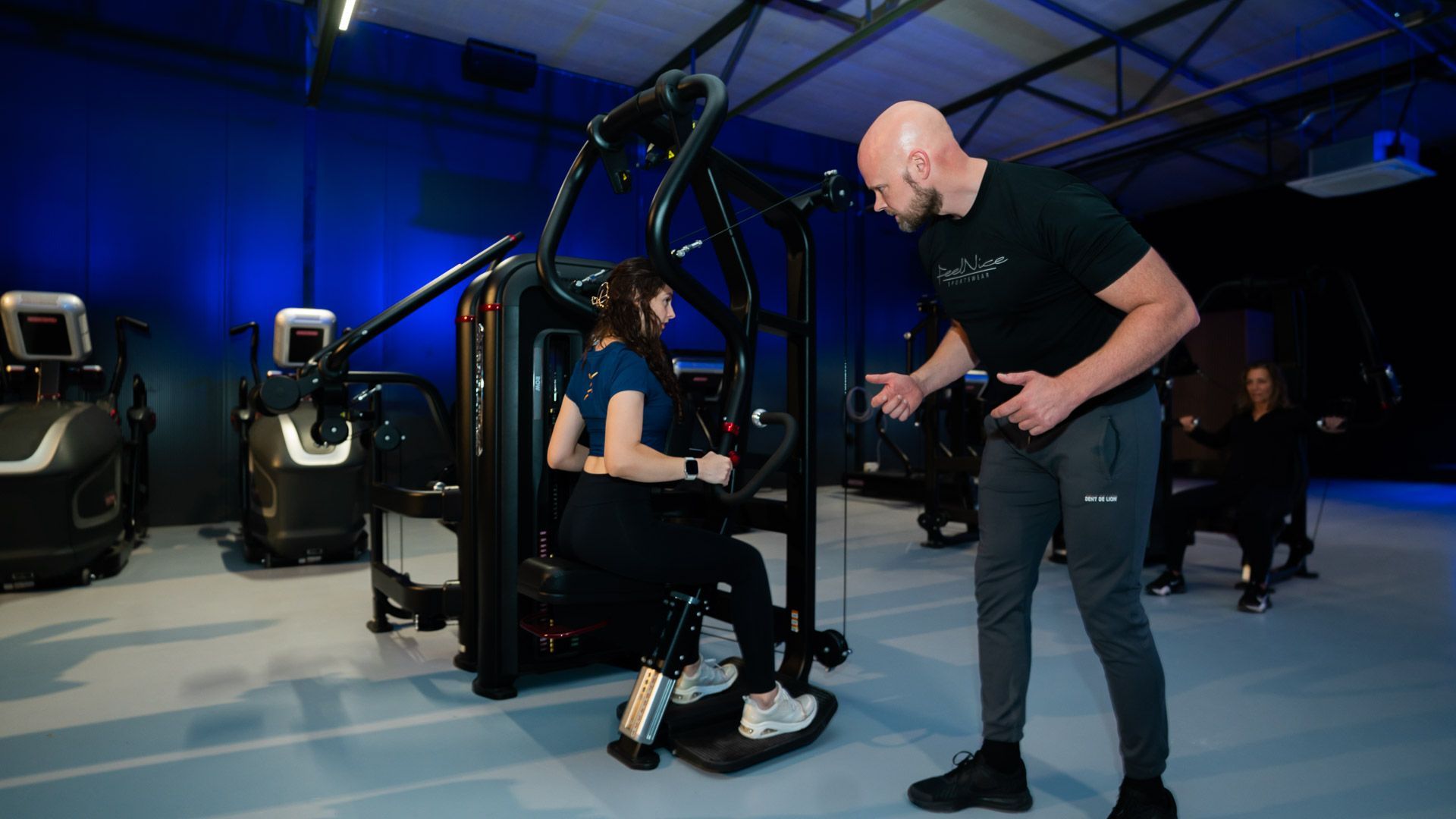 Man in sportkleding rust op een gewichtsapparaat in een sportschool, met een fles water in zijn handen.