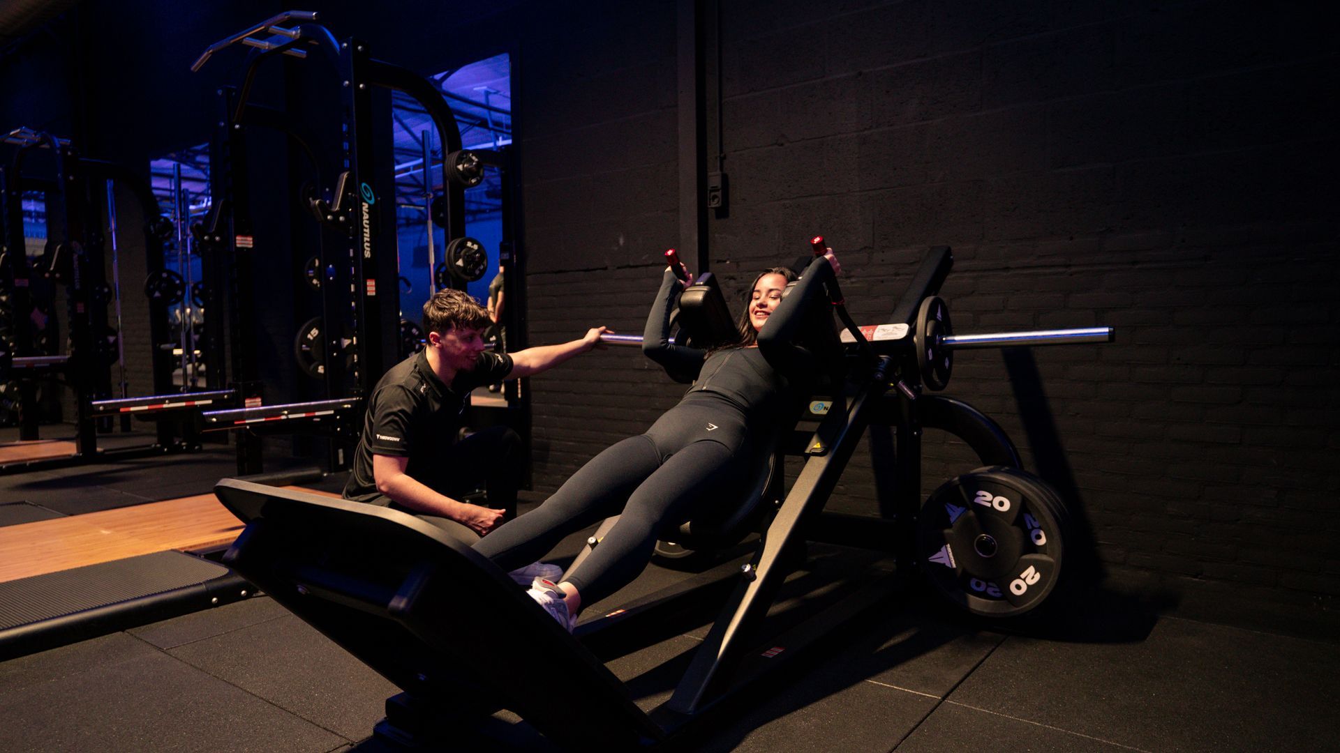Vrouw tilt een halter op met hulp van een trainer in de sportschool.