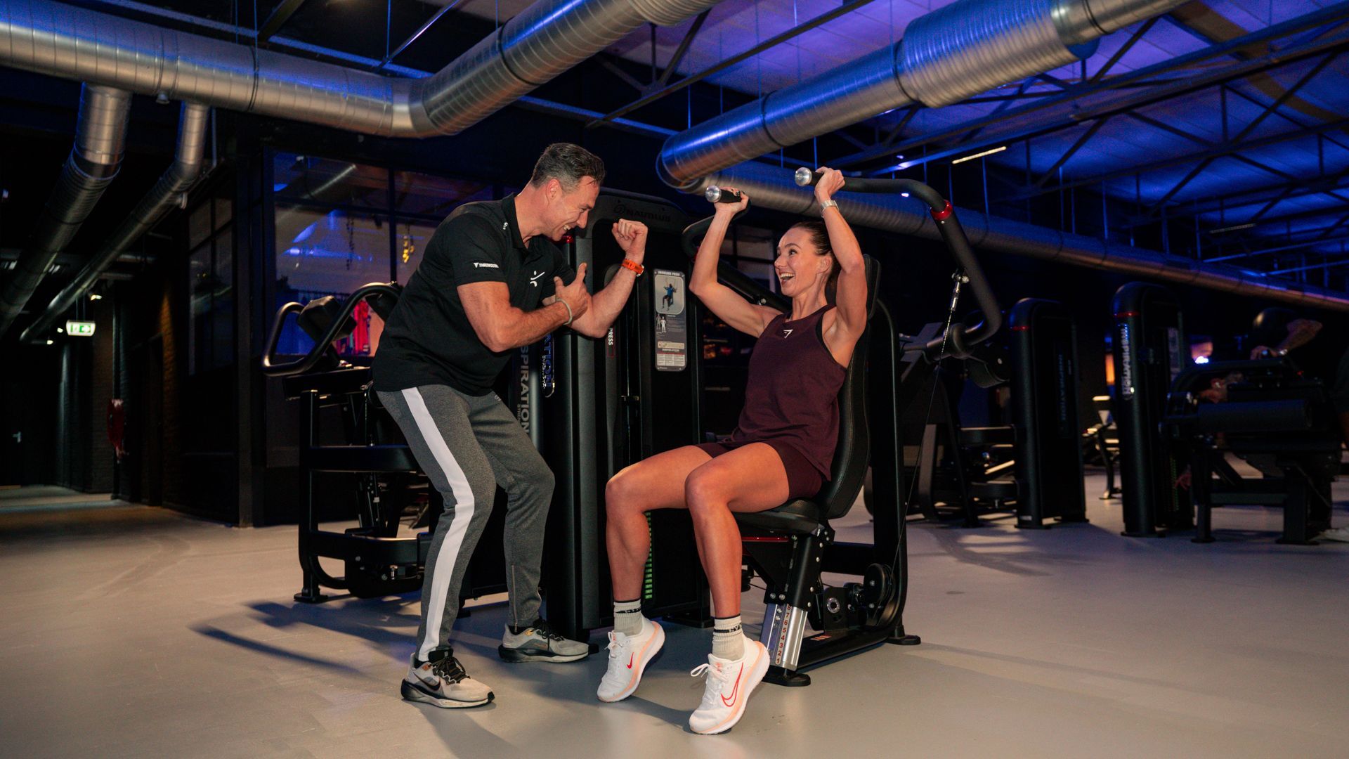 Vrouw in de sportschool, gebogen over dumbbell row op een bankje, draagt een rode legging en een wit topje.
