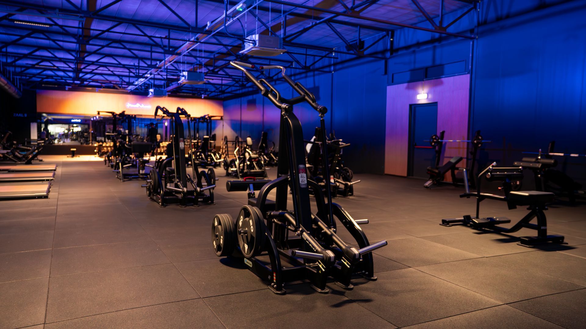 Man in sportkleding rust op een gewichtsapparaat in een sportschool, met een fles water in zijn handen.