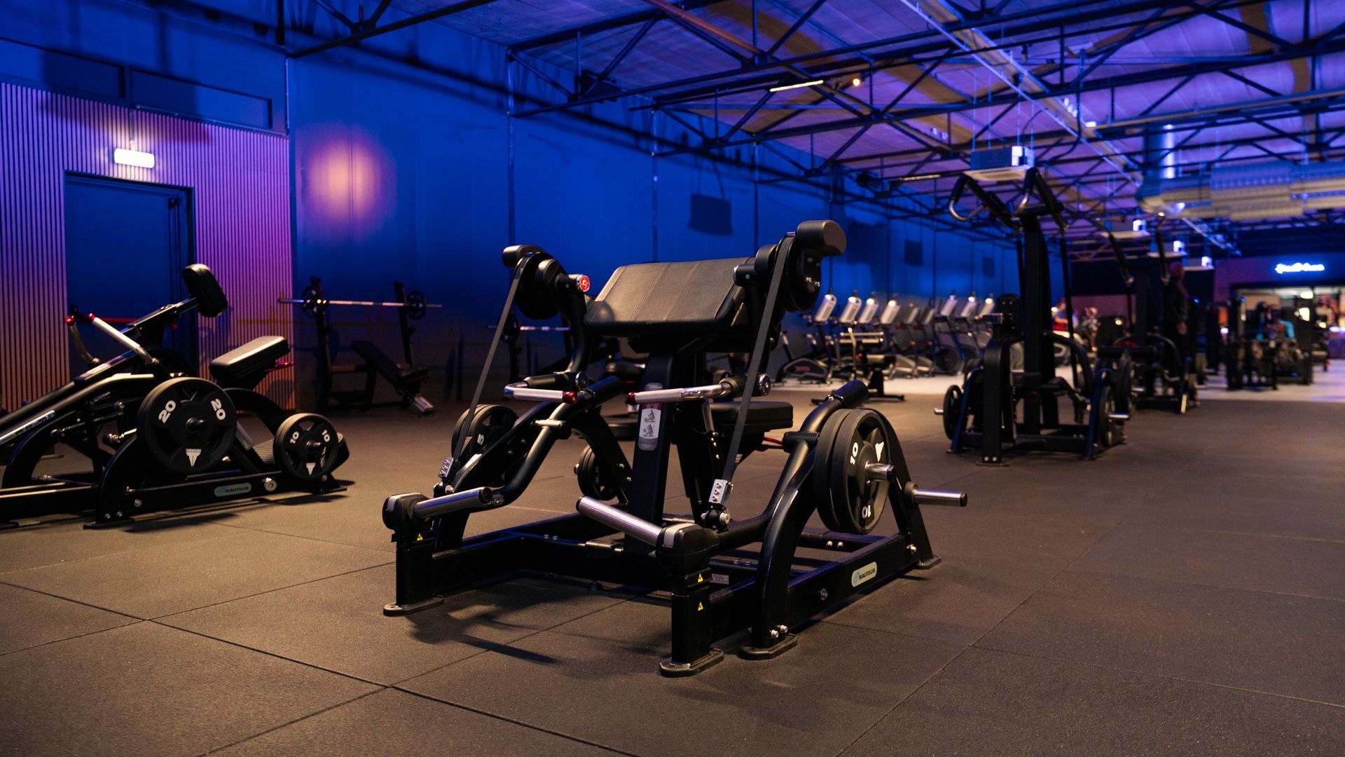 Man in sportschool met halter en gebogen arm. Draagt een neongroen shirt. Achtergrond: fitnessapparatuur, muurschildering in bos.