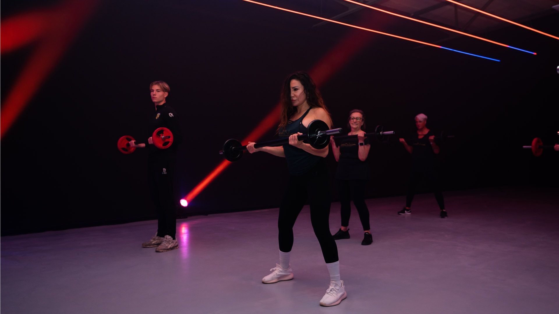 Mensen oefenen met gewichten in een sportschool; vrouw in een roze tanktop lacht terwijl ze een kettlebell tilt.