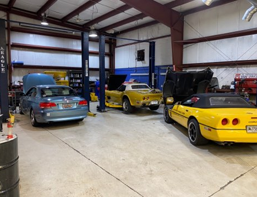 Classic cars in a workshop garage, including a blue sedan and two yellow sports cars on lifts.