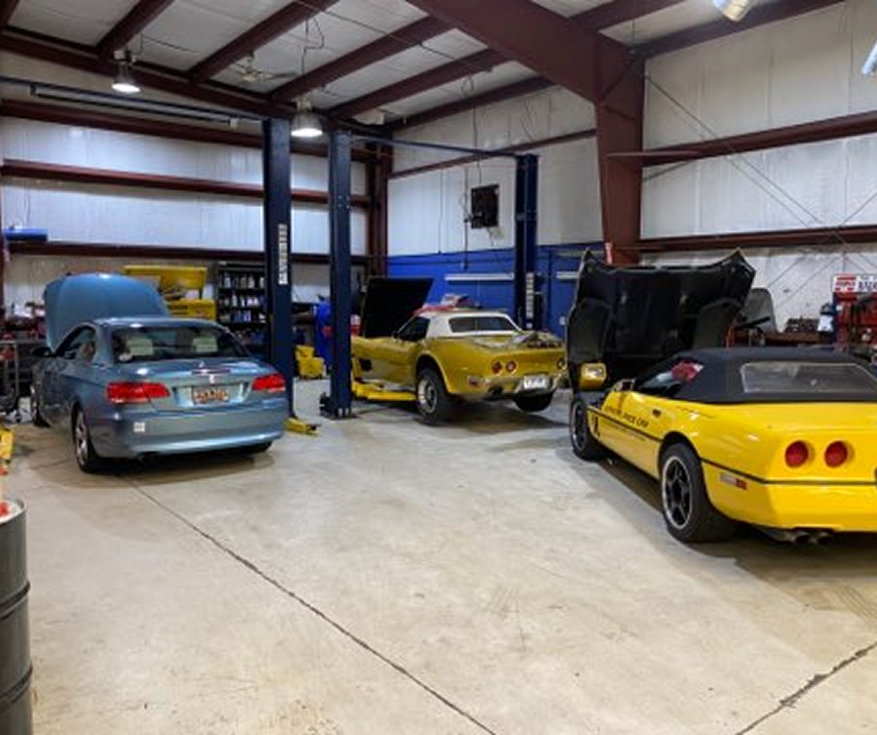 Classic cars in a workshop garage, including a blue sedan and two yellow sports cars on lifts.