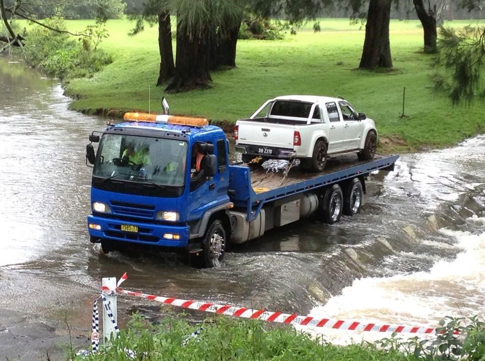 A blue tow truck is towing a white ute across a bridge with water — Quick Hitch Towing & Tilt Tray Services Illawarra in Oak Flats, NSW