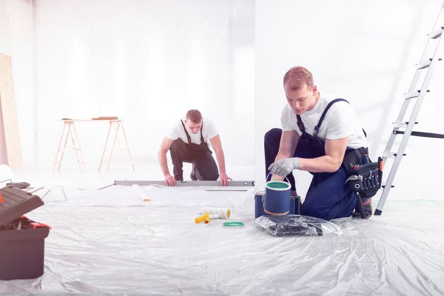 Two workers in blue overalls painting a room. One kneels with paint supplies, the other measures the floor.