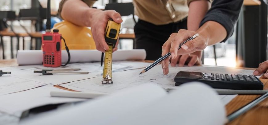 People inspecting blueprints with a measuring tape, calculator, and pencil. Yellow hard hat and walkie-talkie on desk.