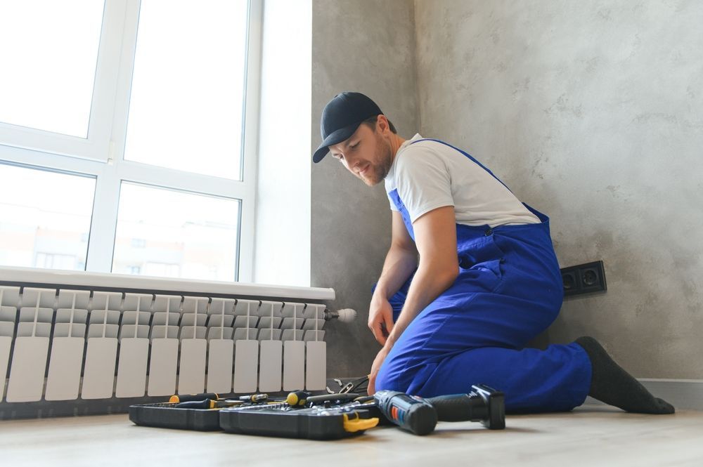 A person in blue overalls kneels, working on wiring near a window and radiator. Tools are in a tray.
