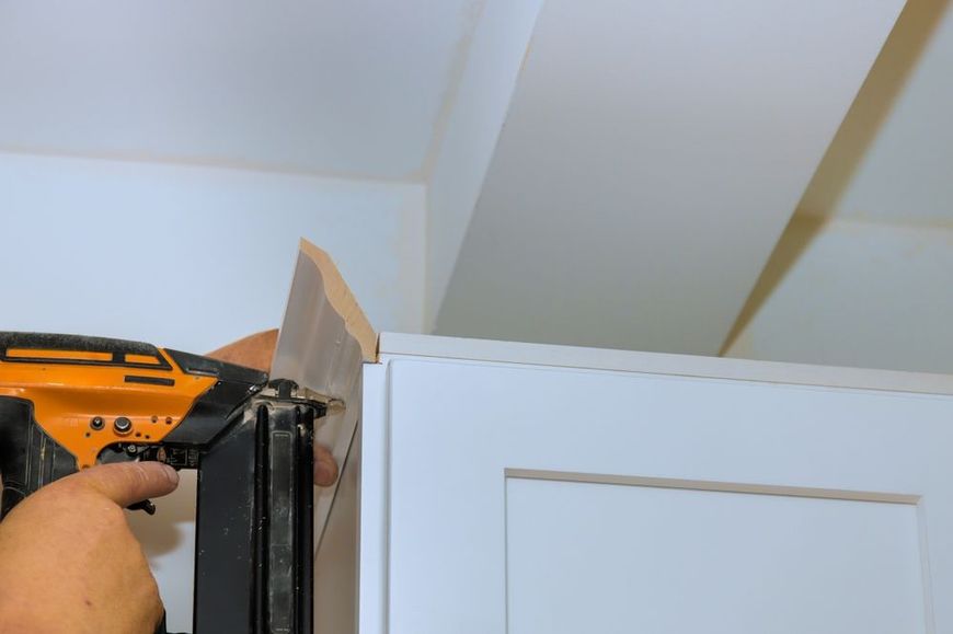 A person using a nail gun to install trim on a white cabinet.