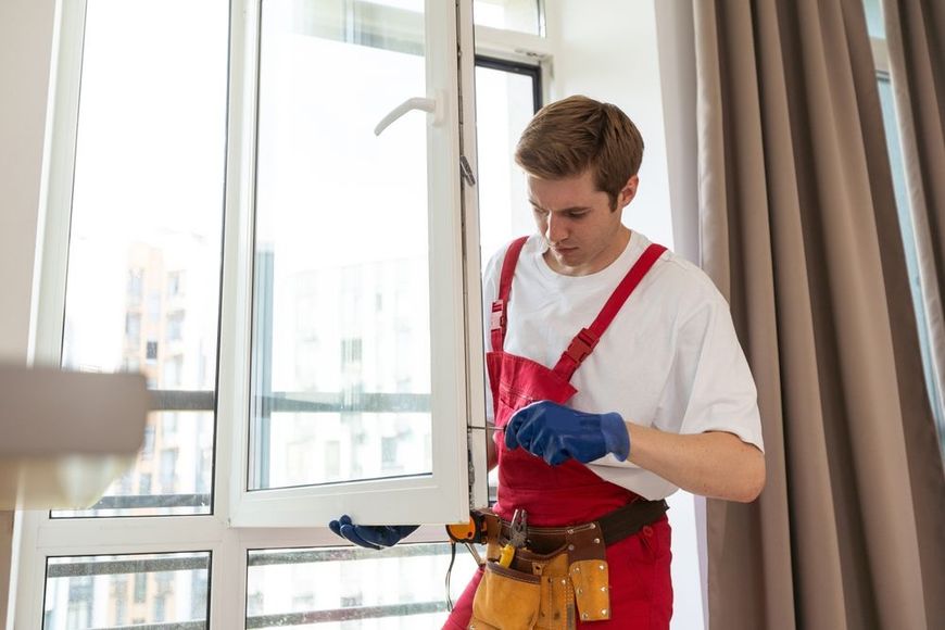 Man in red overalls and blue gloves working on a window with a tool belt.