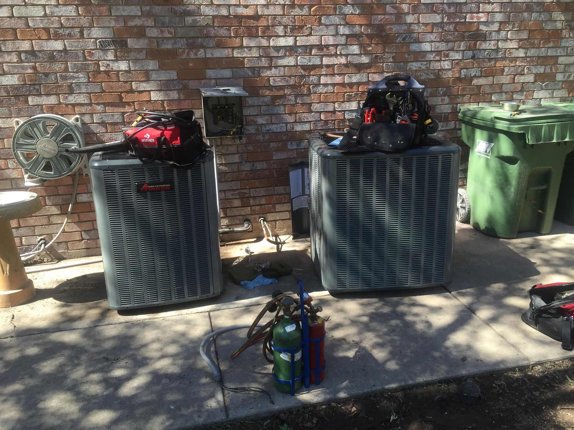 Two air conditioners are sitting on a sidewalk in front of a brick wall.