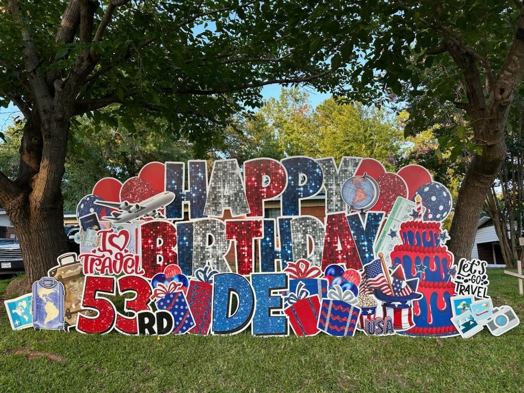 A large happy birthday sign is sitting in the grass in front of a tree.