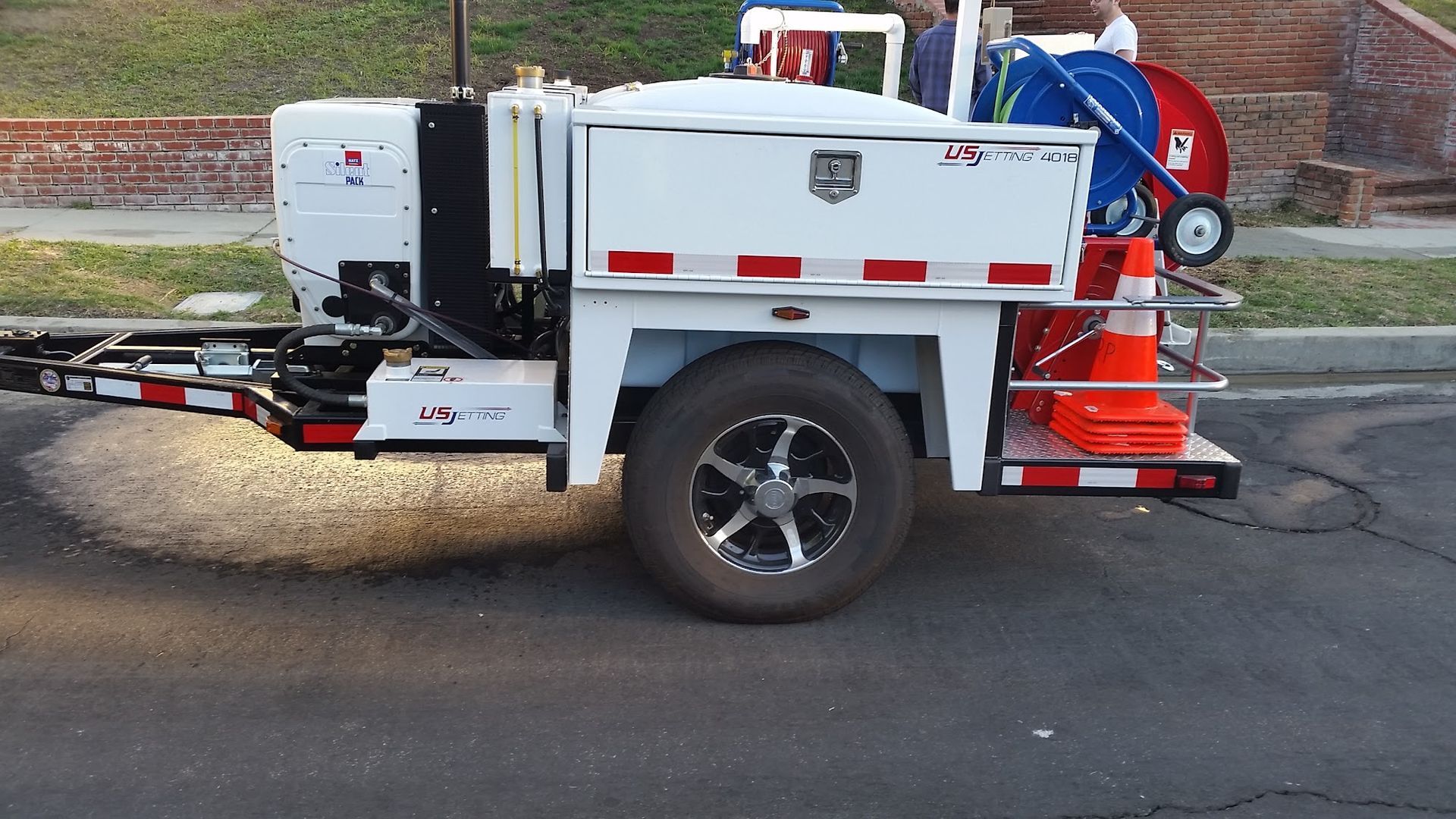 White utility trailer with equipment on street. Red and white reflective tape, orange traffic cone, and black tire visible.