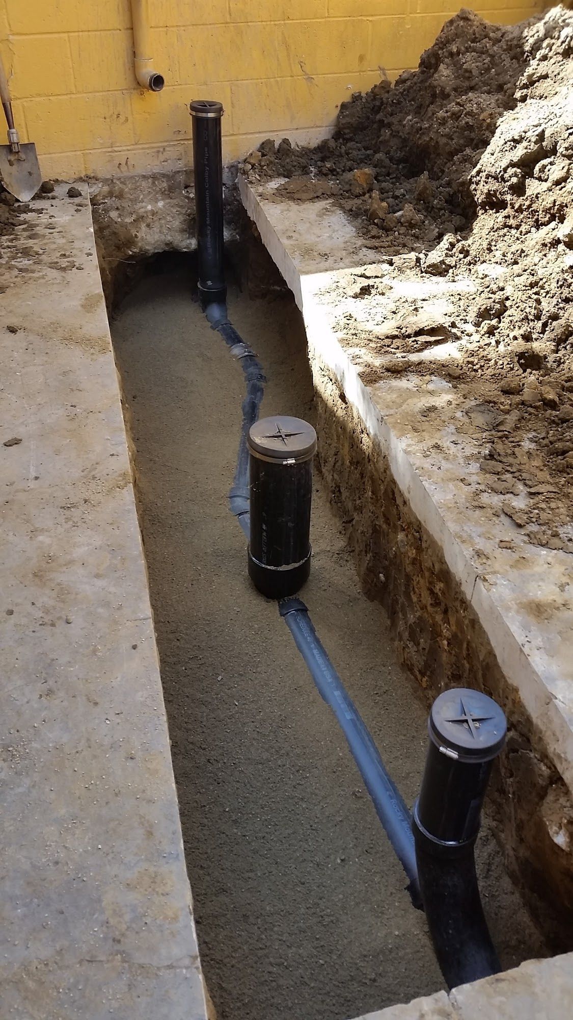 Trench with black pipes and vertical capped tubes, set in gravel against a yellow wall and soil.