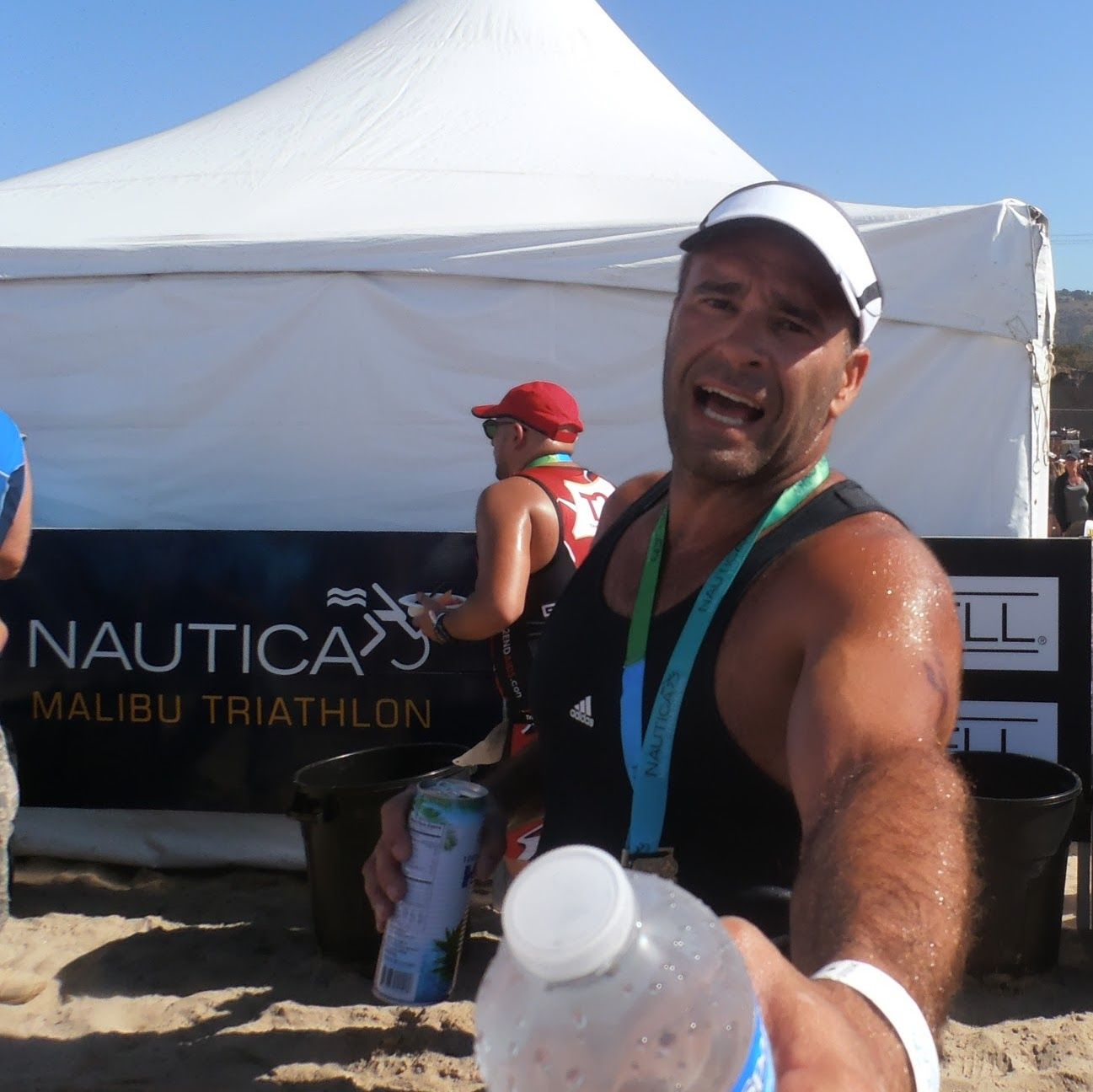 Man at Malibu Triathlon, sweaty, holding water bottle, white tent in background.