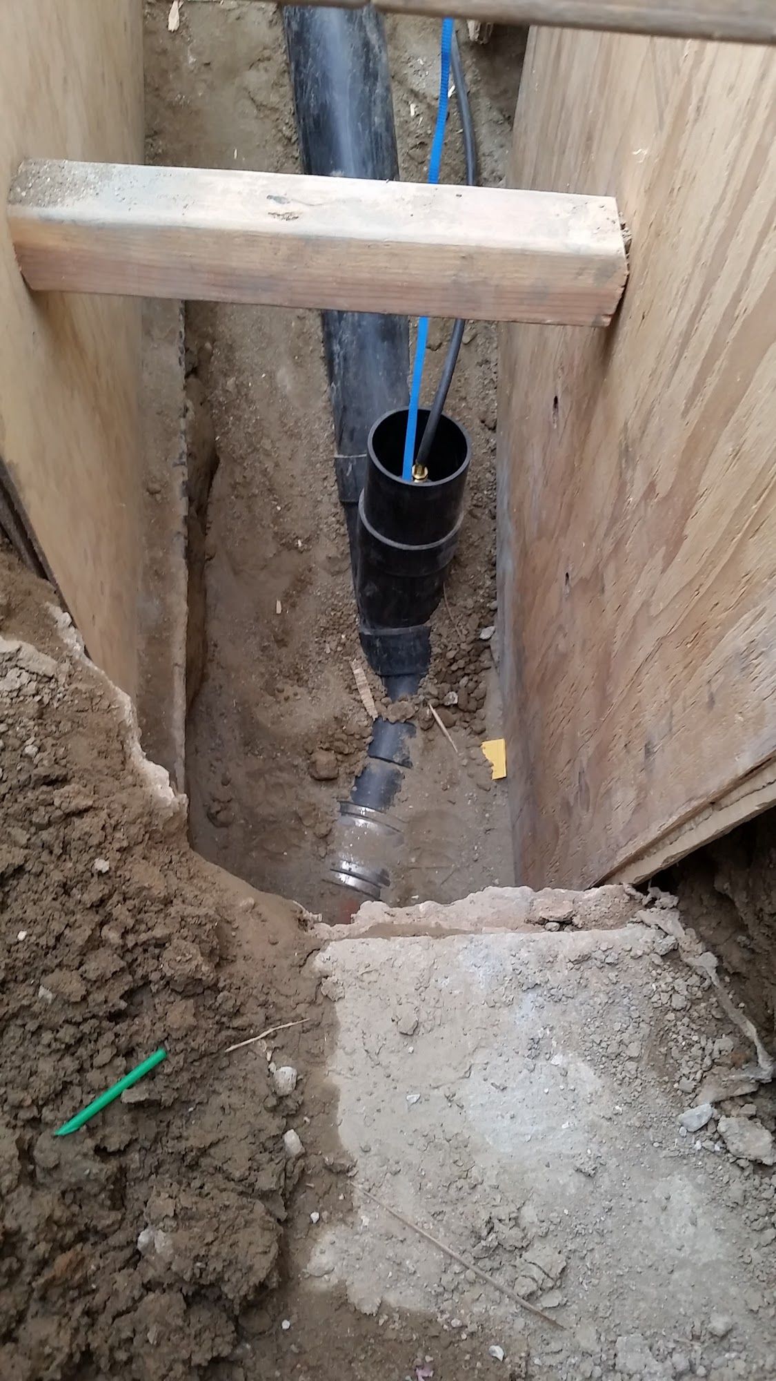 Trench with black PVC pipes and blue water lines, wood support beams and plywood walls.