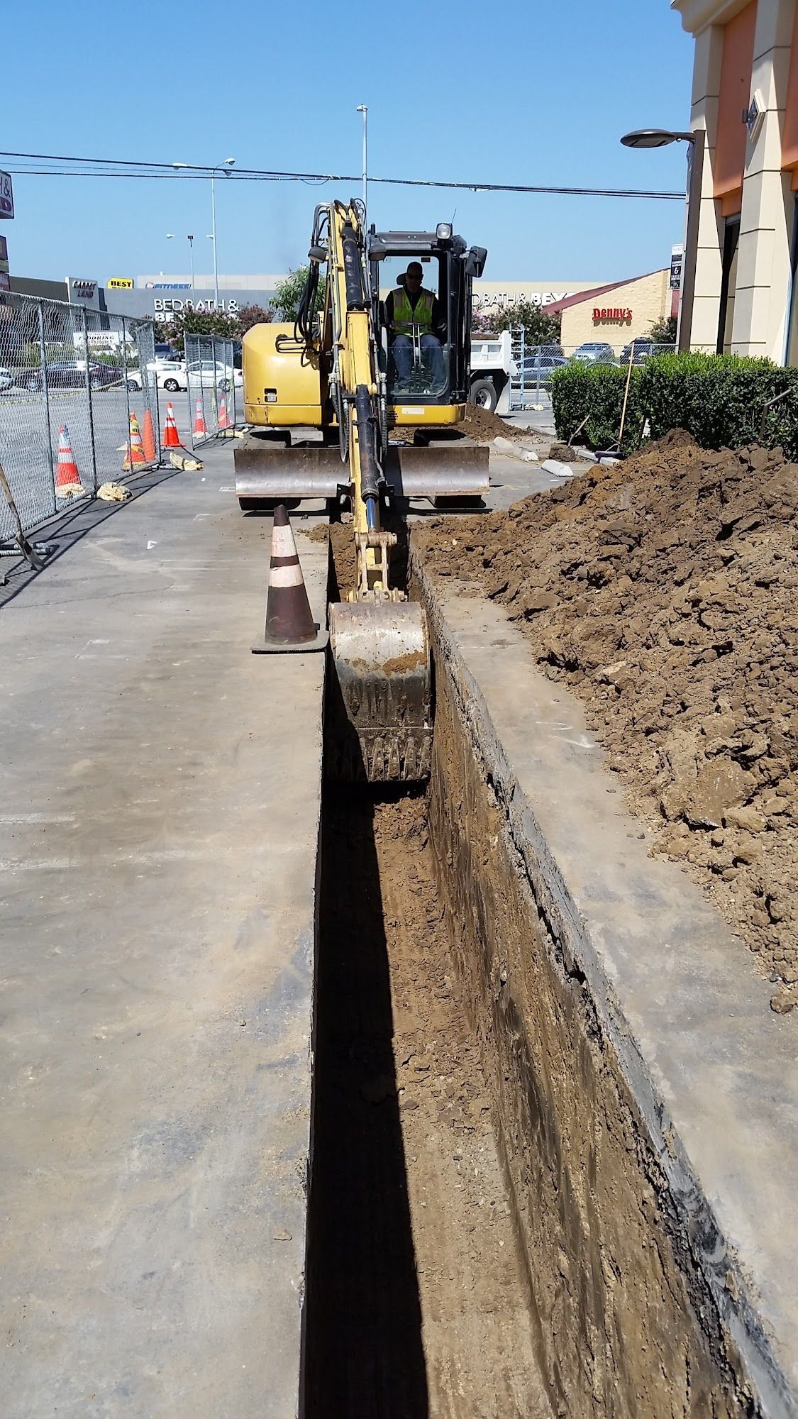 Yellow excavator digging a long trench along a concrete sidewalk next to a road.