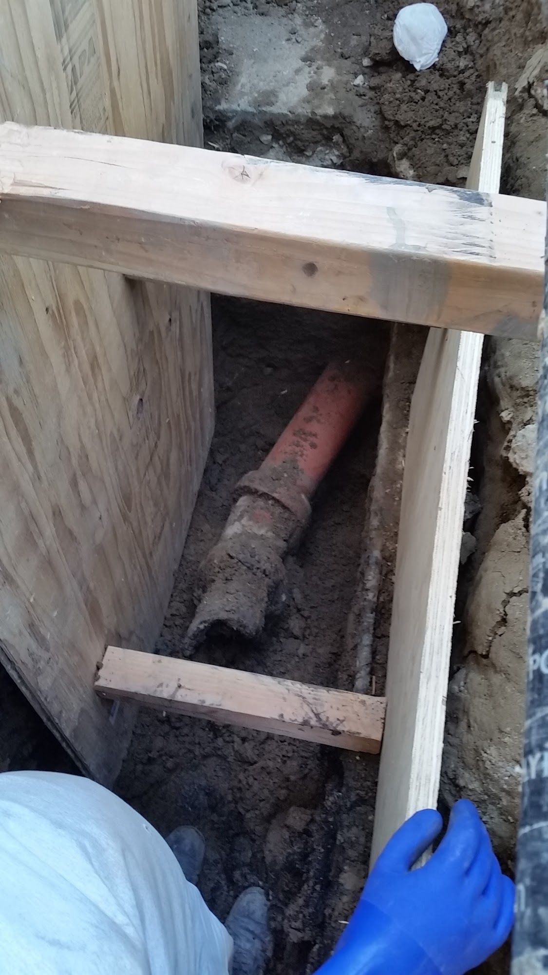 Orange pipe in earthen trench, supported by wooden bracing. Blue-gloved hand visible.