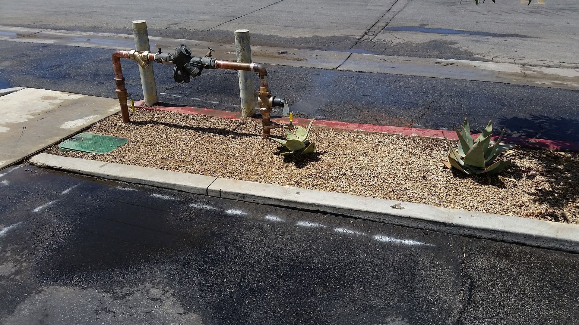 Copper pipes and valves, gravel bed with plants, next to asphalt pavement.