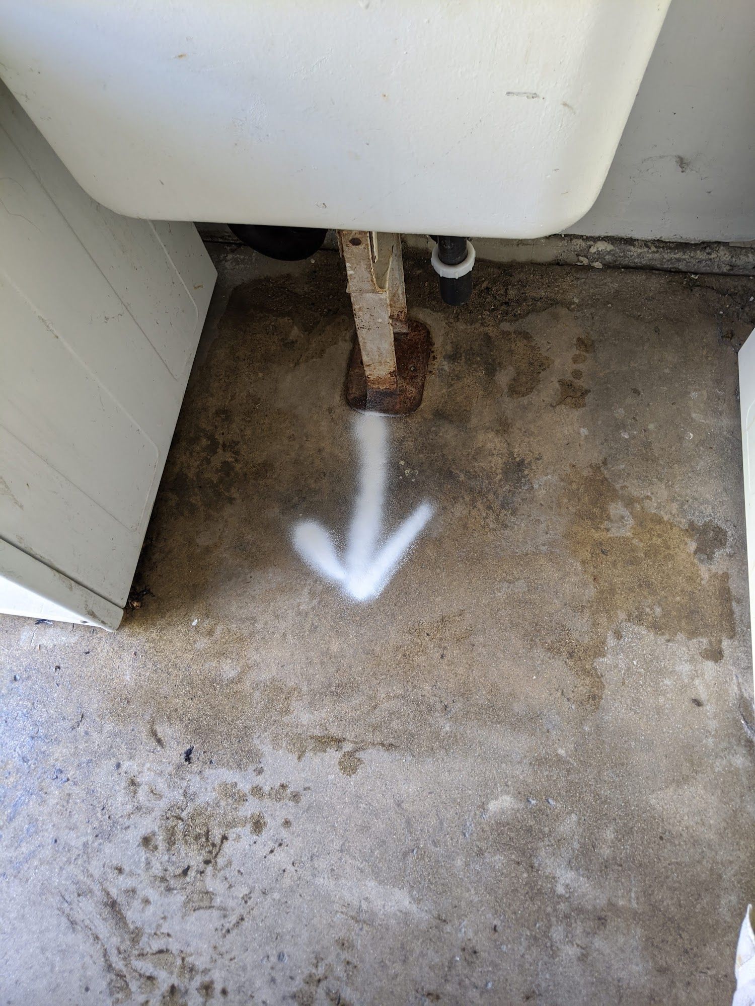 White arrow spray-painted on a concrete floor points toward a rusty pipe beneath a white sink.