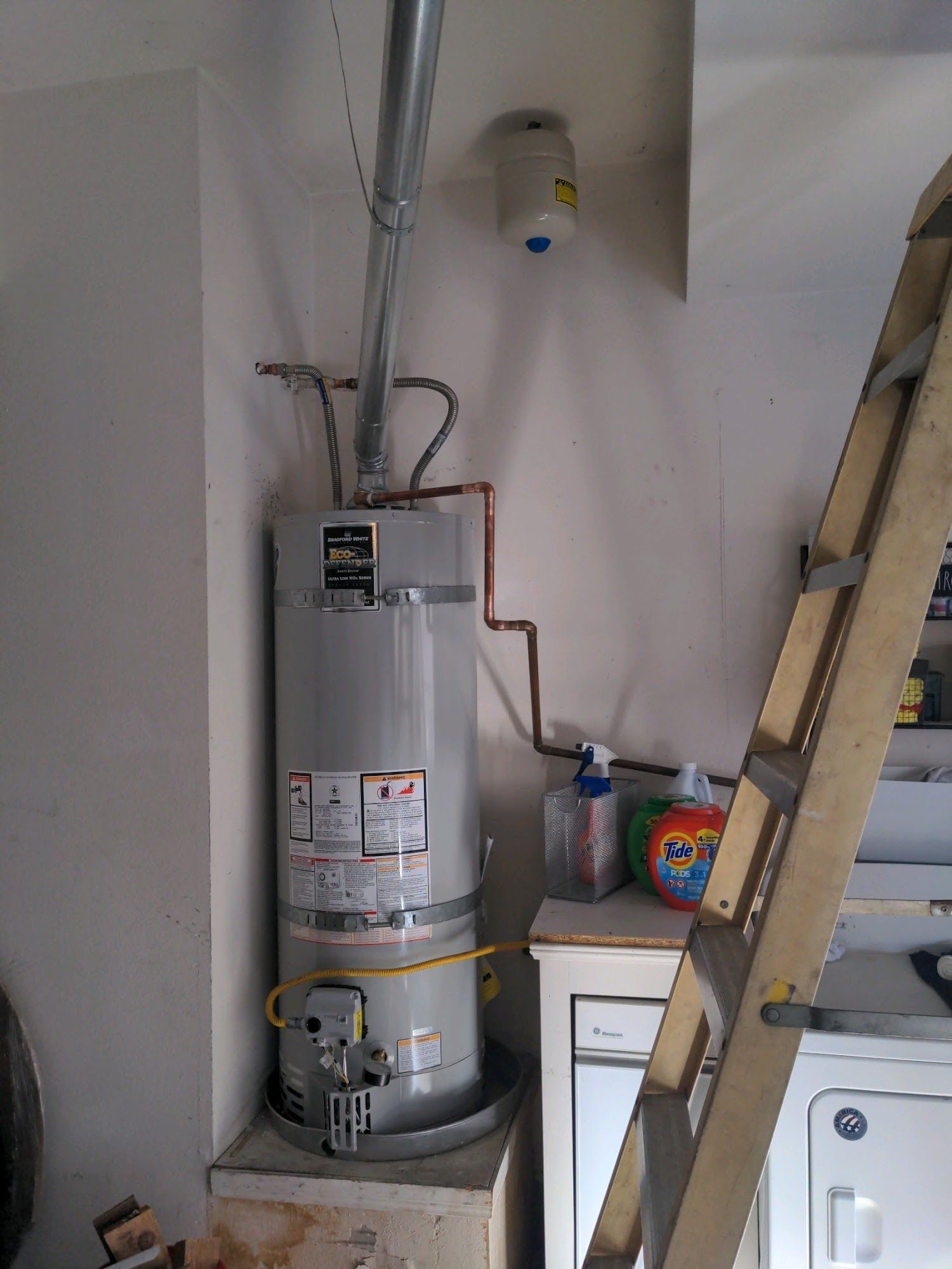 Tall gray water heater in a garage, next to a ladder and laundry appliances.