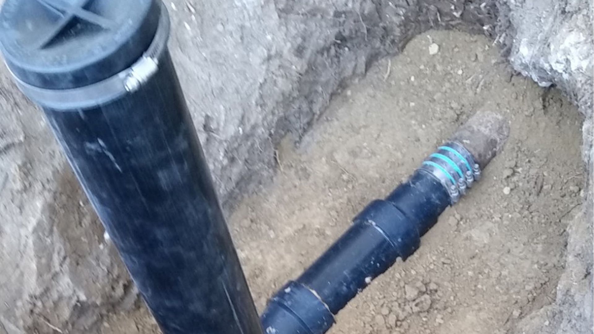 Black pipes and fittings in a dirt hole, possibly underground plumbing repair.