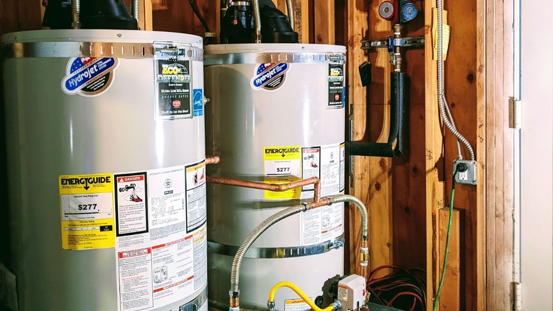 Two water heaters side-by-side in a wooden utility room. Copper and black pipes connect them to other equipment.