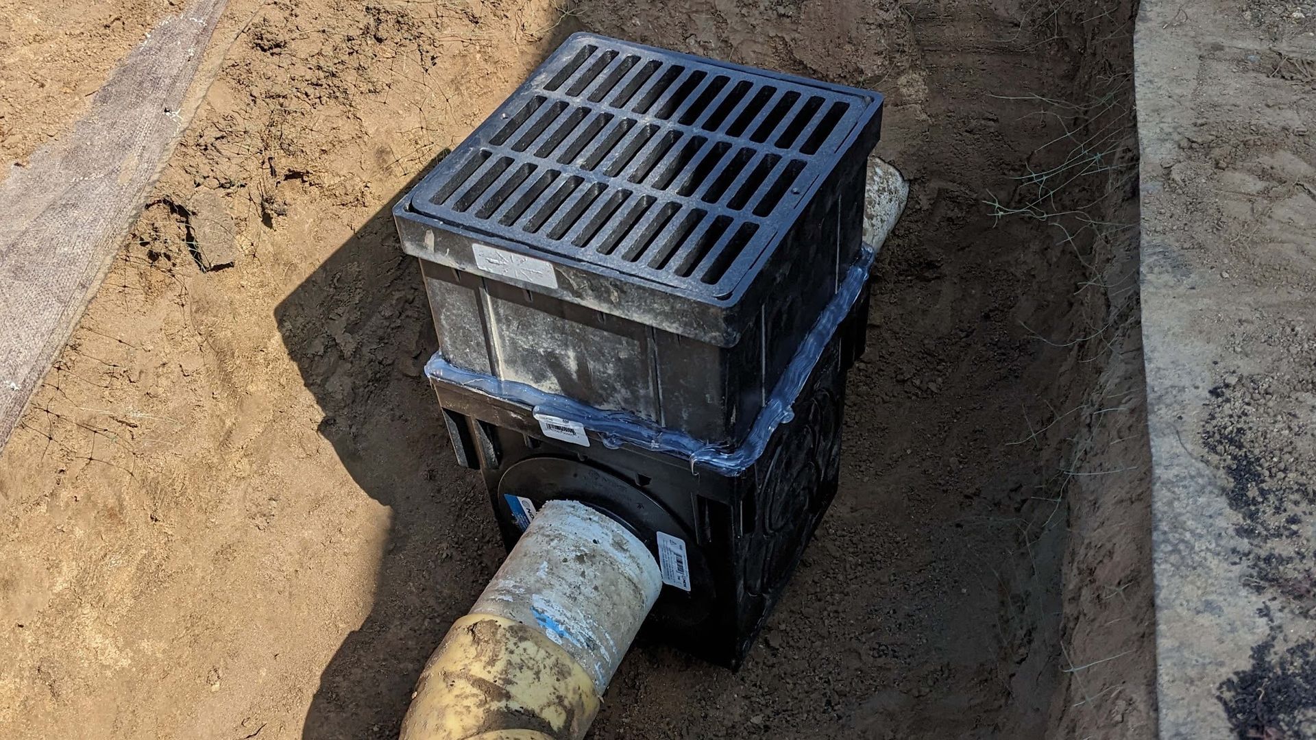 Black stormwater drain with grate installed in a trench, connected to a pipe.