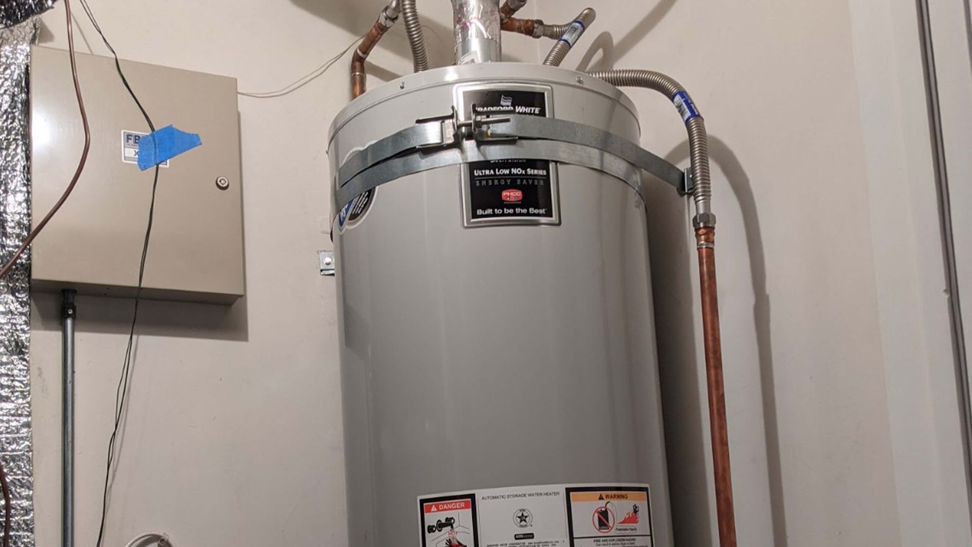 A water heater with metal bands and copper pipes in an indoor utility room, next to a metal box.