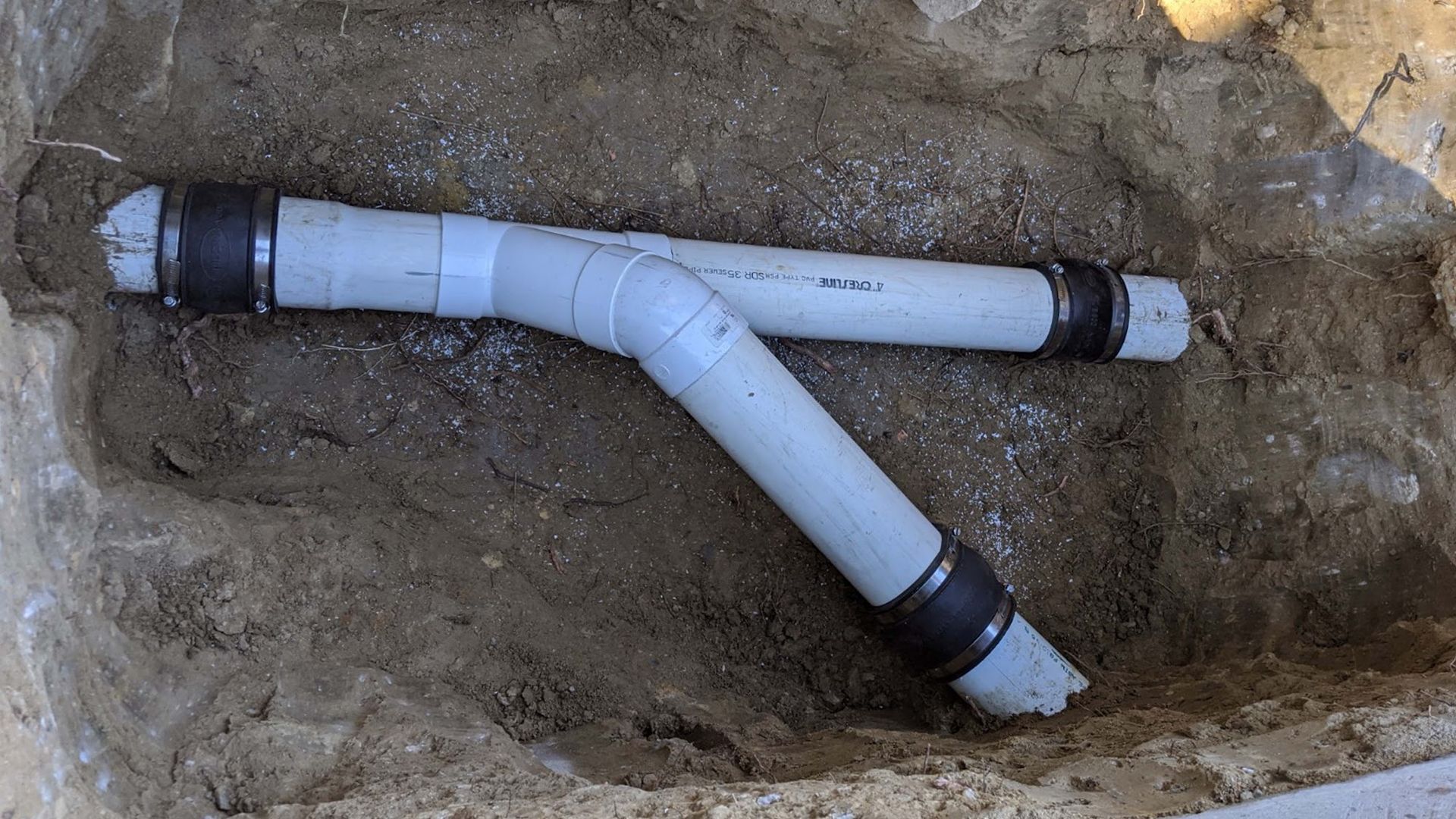 White PVC pipes with black couplings in a dirt trench, forming a Y-shape.