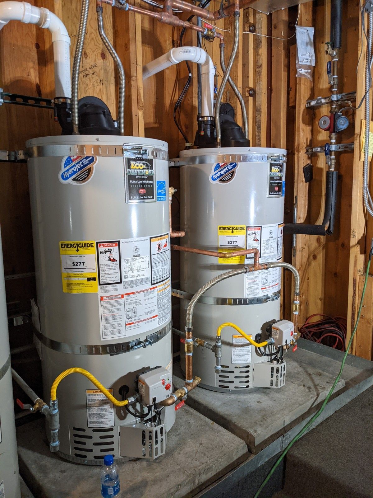 Two gas water heaters side-by-side in a utility room. Yellow gas lines and copper pipes are visible.