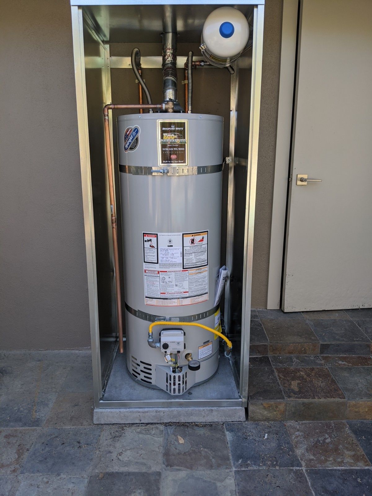 A gas water heater enclosed in a metal frame, mounted on a stone patio next to a door.