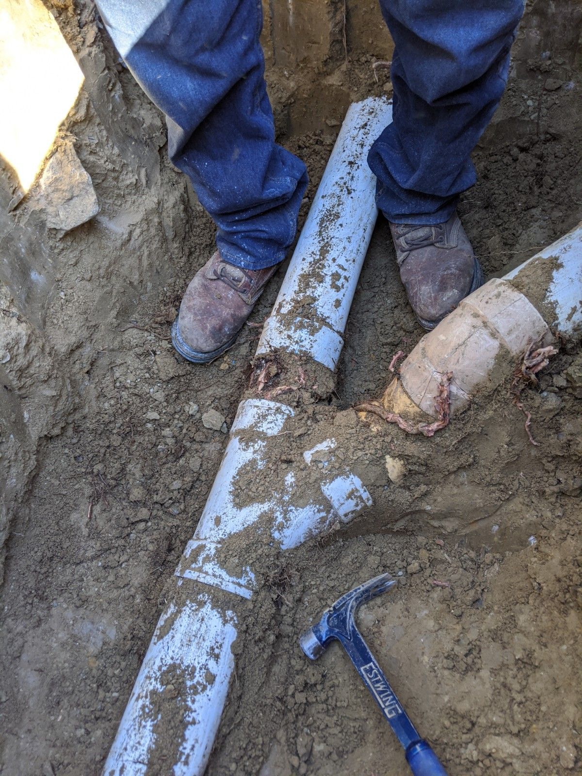 Person repairing broken pipes in a trench, using a hammer, surrounded by dirt.