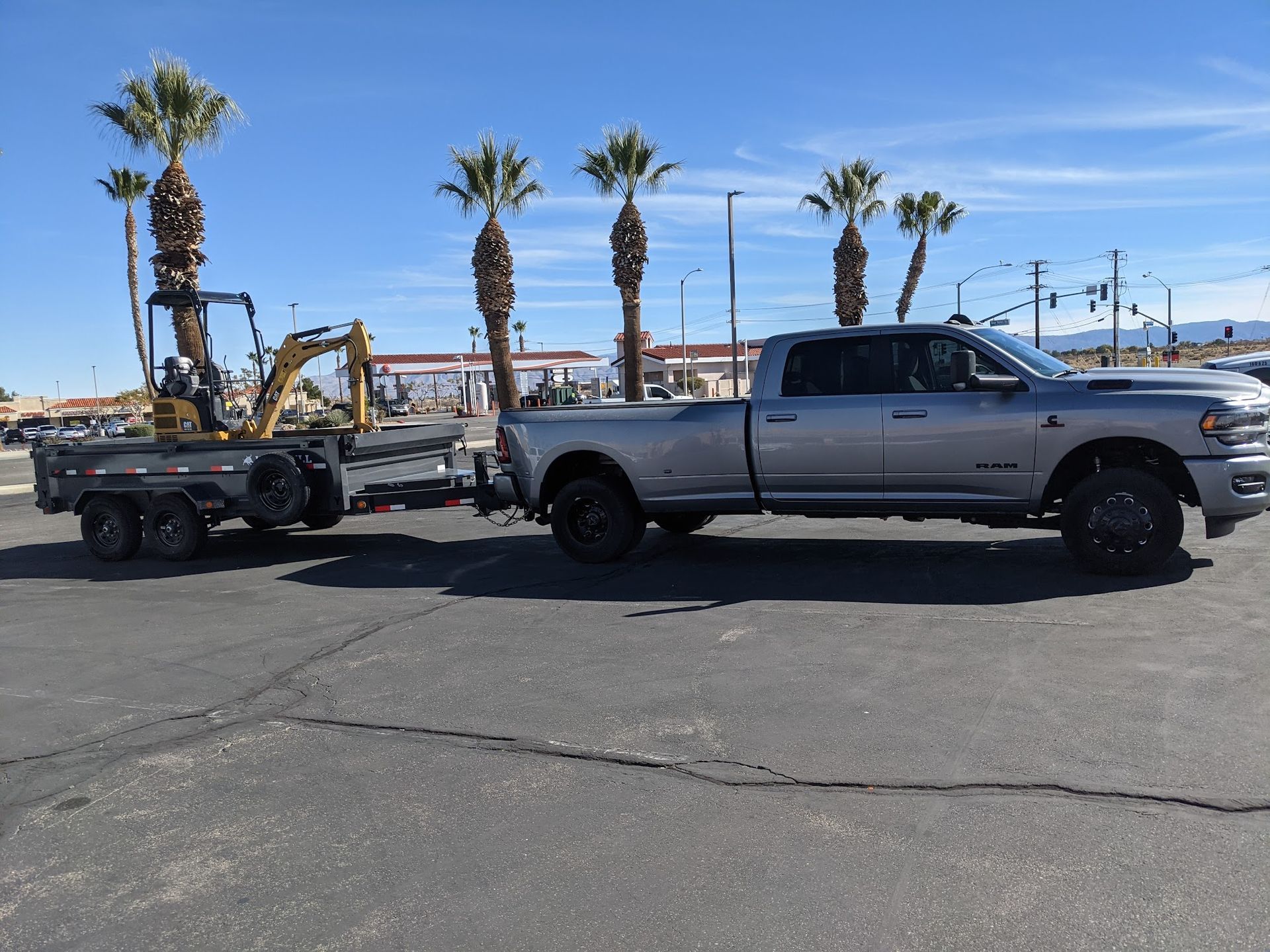 Gray pickup truck towing a trailer with a small excavator on a paved lot; palm trees in the background.