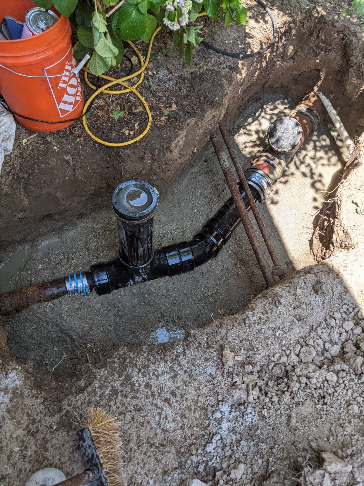 A sprinkler system repair: a pipe connection in a trench, with a Home Depot bucket and tools nearby.