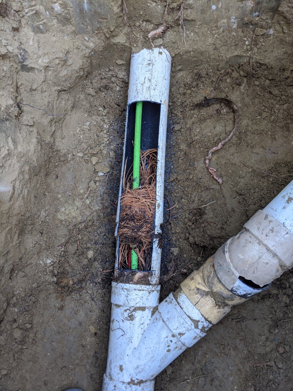 Broken white PVC pipe in a dirt trench, filled with roots, showing a green pipe inside.