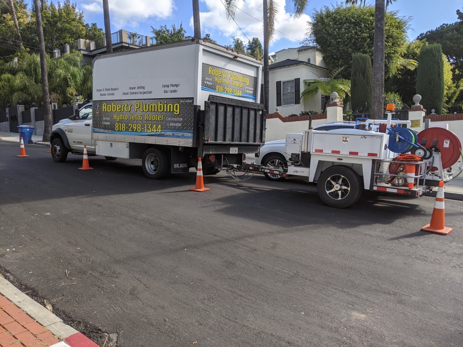 Large truck with open dump bed and trailer on a newly paved street, next to orange traffic cones.