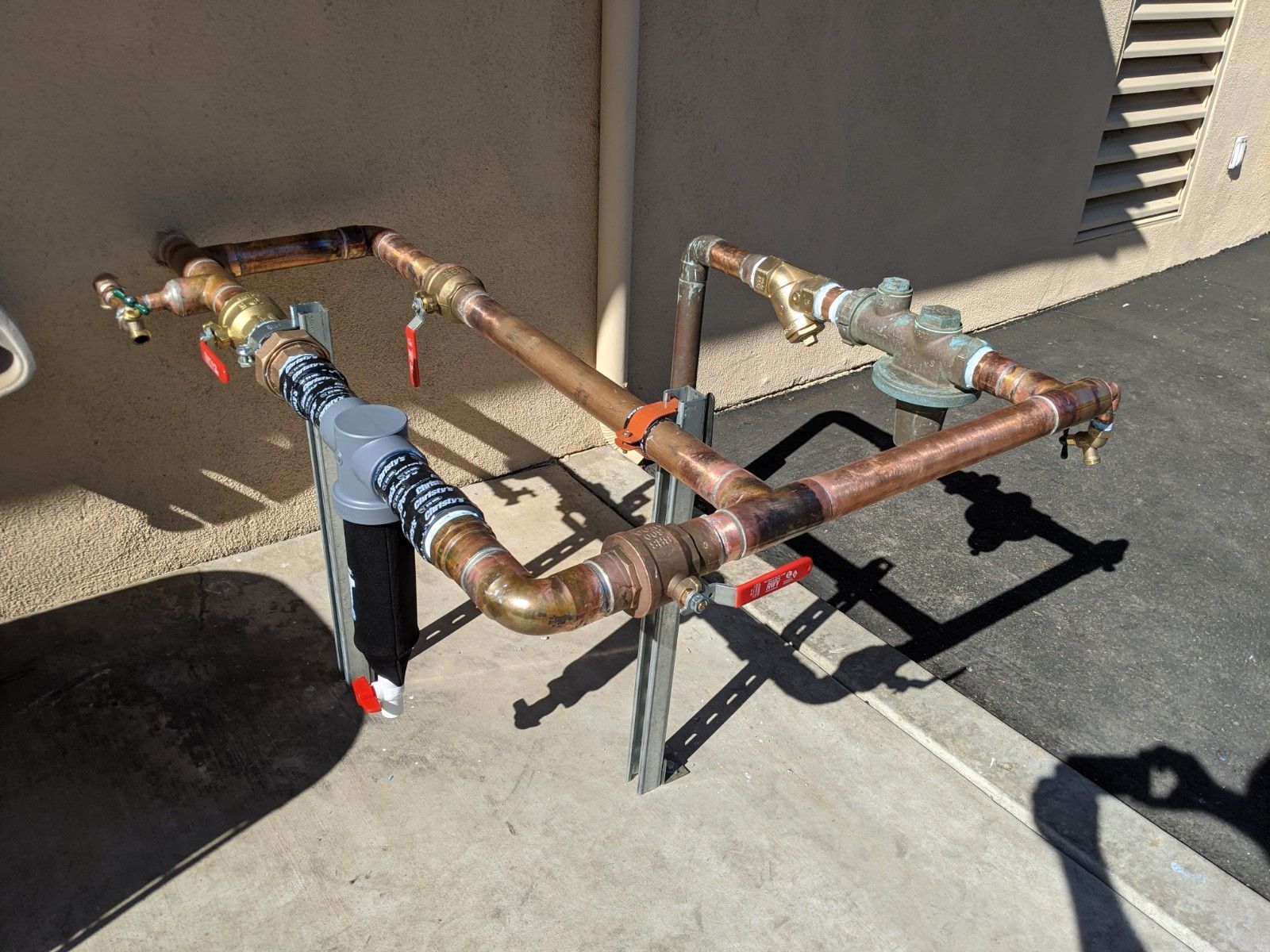 Copper water pipes with valves and a filter outside on a concrete surface.
