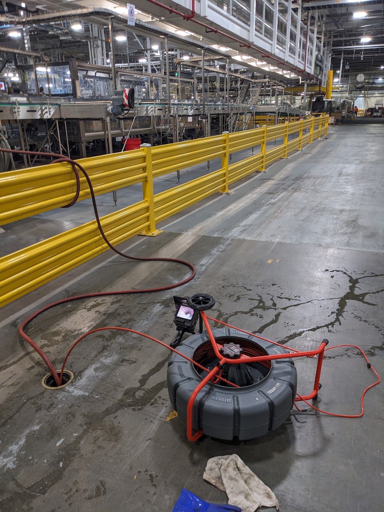 A gray hose reel with red hoses on a factory floor, yellow barriers and machinery in the background.