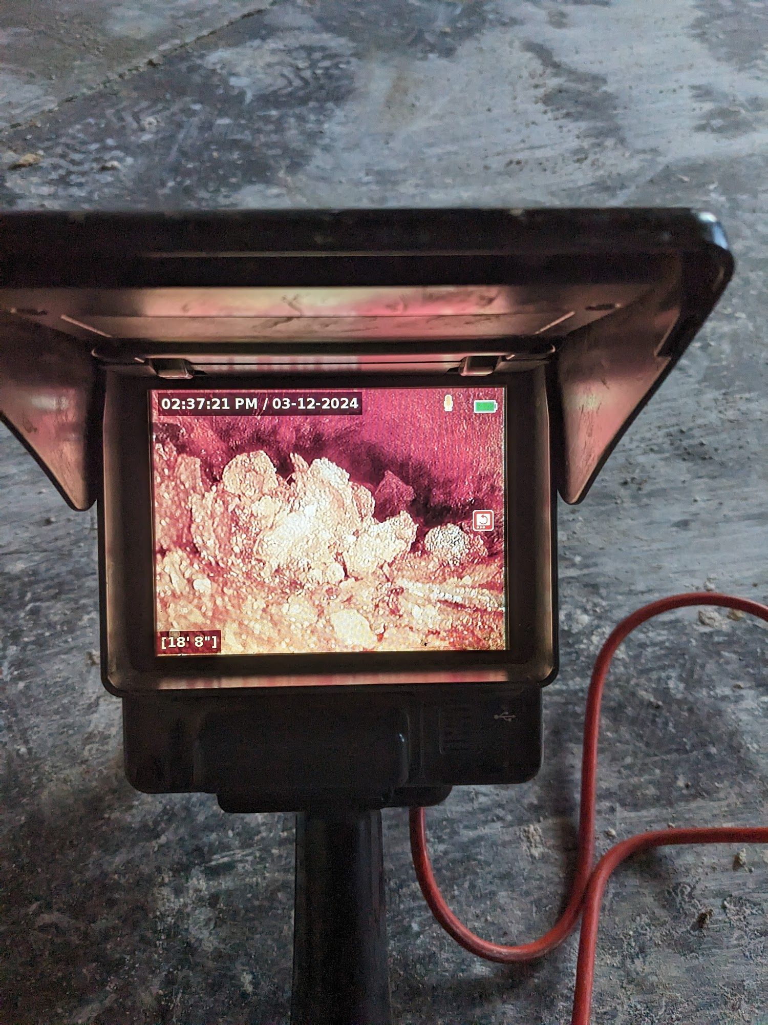 Thermal camera display showing a dark red scene with bright white areas, enclosed in a black housing. Red cable attached.