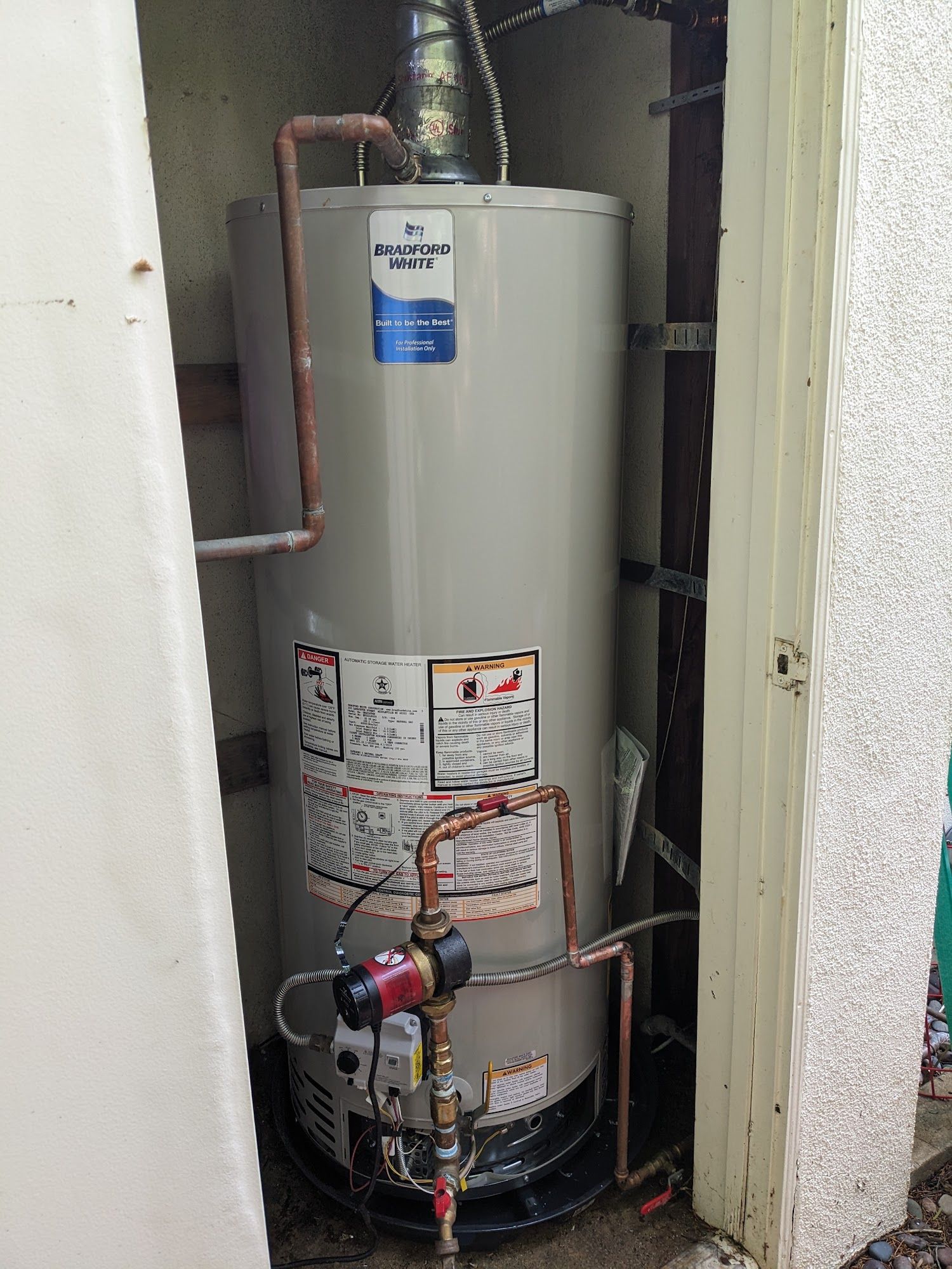A natural gas water heater unit encased in a narrow outdoor enclosure. Copper pipes and vent are visible.