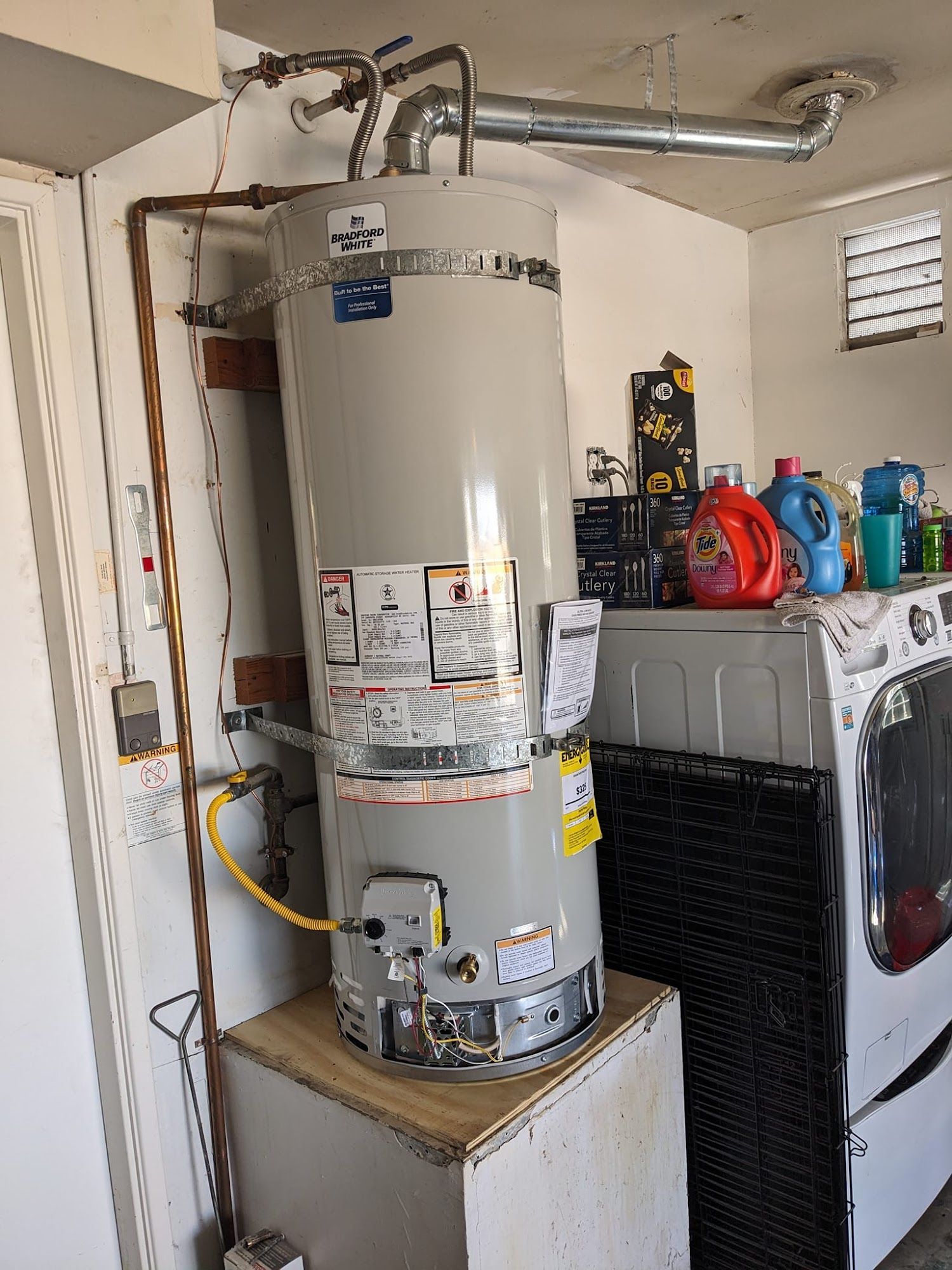 Water heater in a garage, next to a washing machine and storage shelf. Copper pipes and exhaust vent visible.