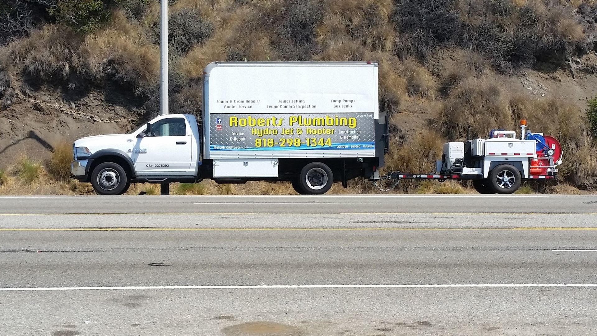 White plumbing truck with trailer parked on roadside; hillside in background.