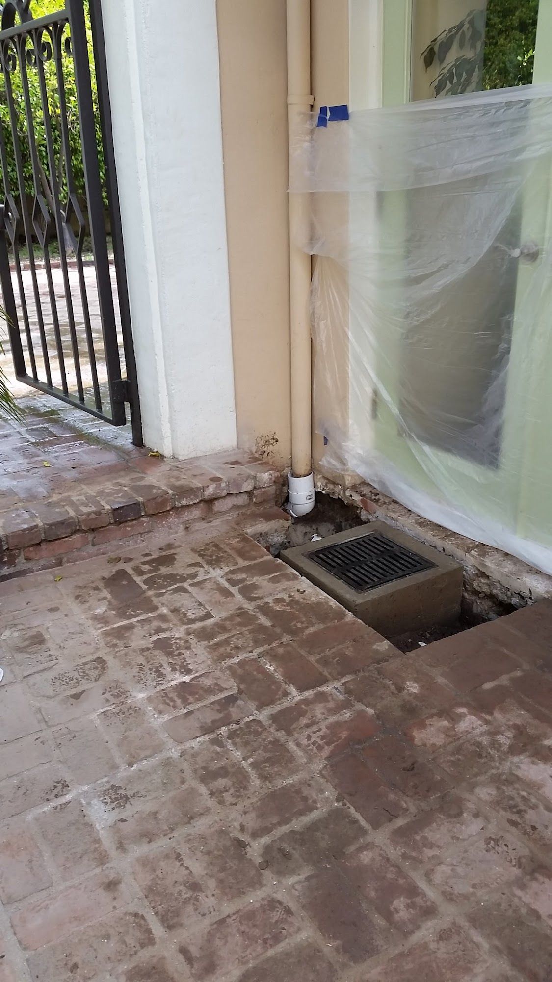 A ground-level view of a concrete drain with a metal grate, next to a beige wall and a plastic-covered door.