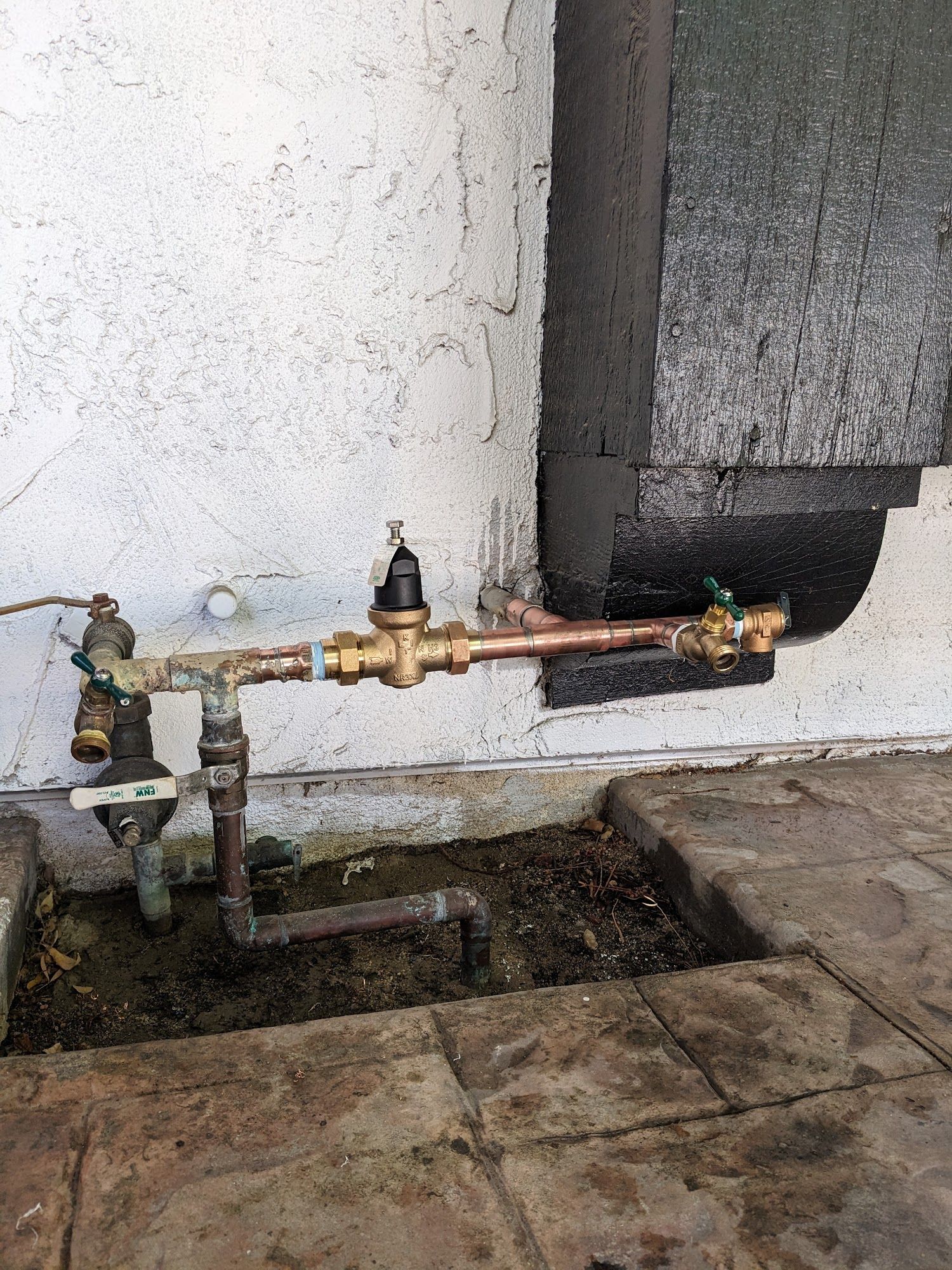 Copper pipes and water valve system attached to a white wall, set in a tiled patio.