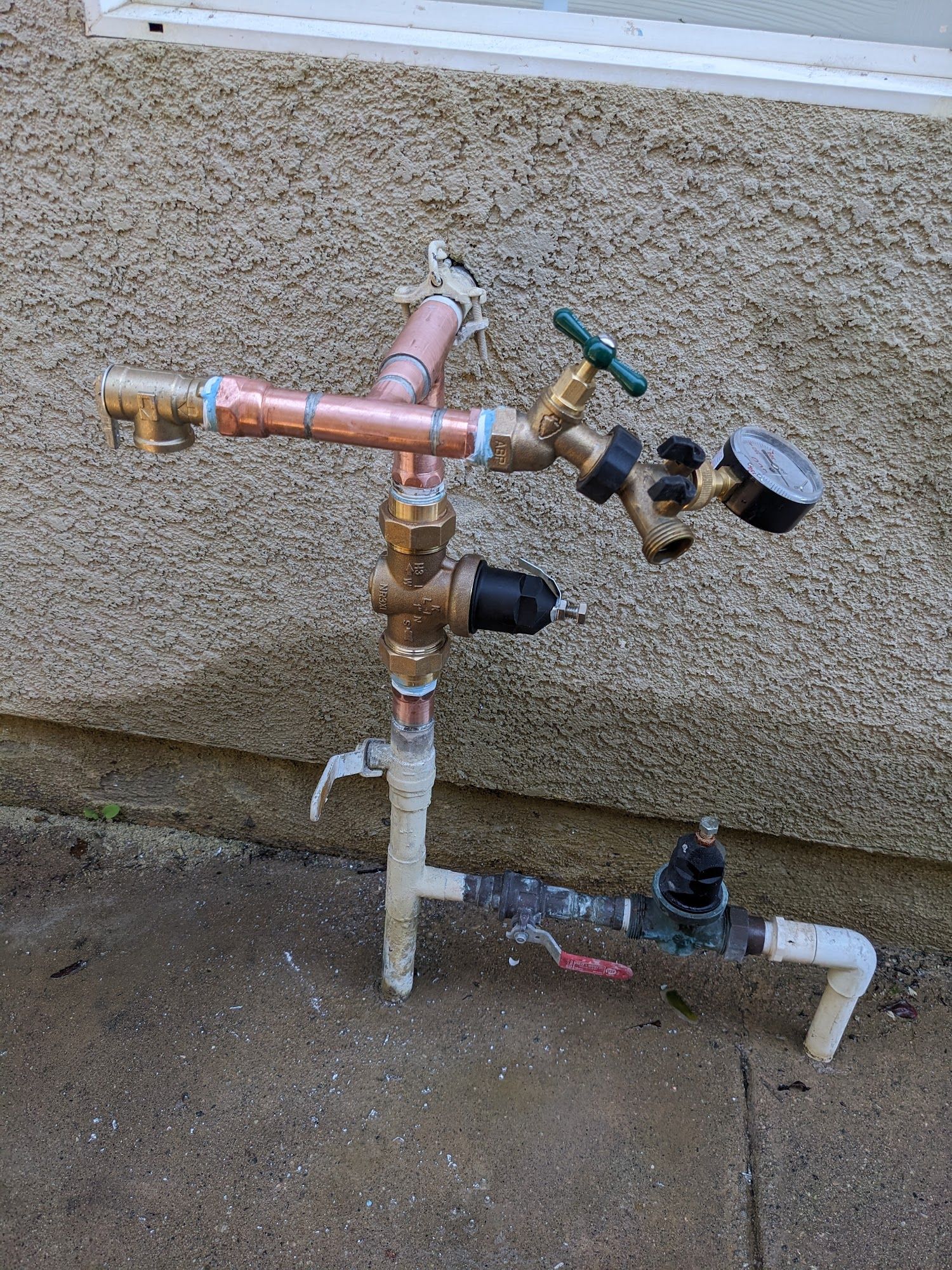 Outdoor water pipe system with multiple valves, a pressure gauge, and copper and PVC pipes on a textured wall.