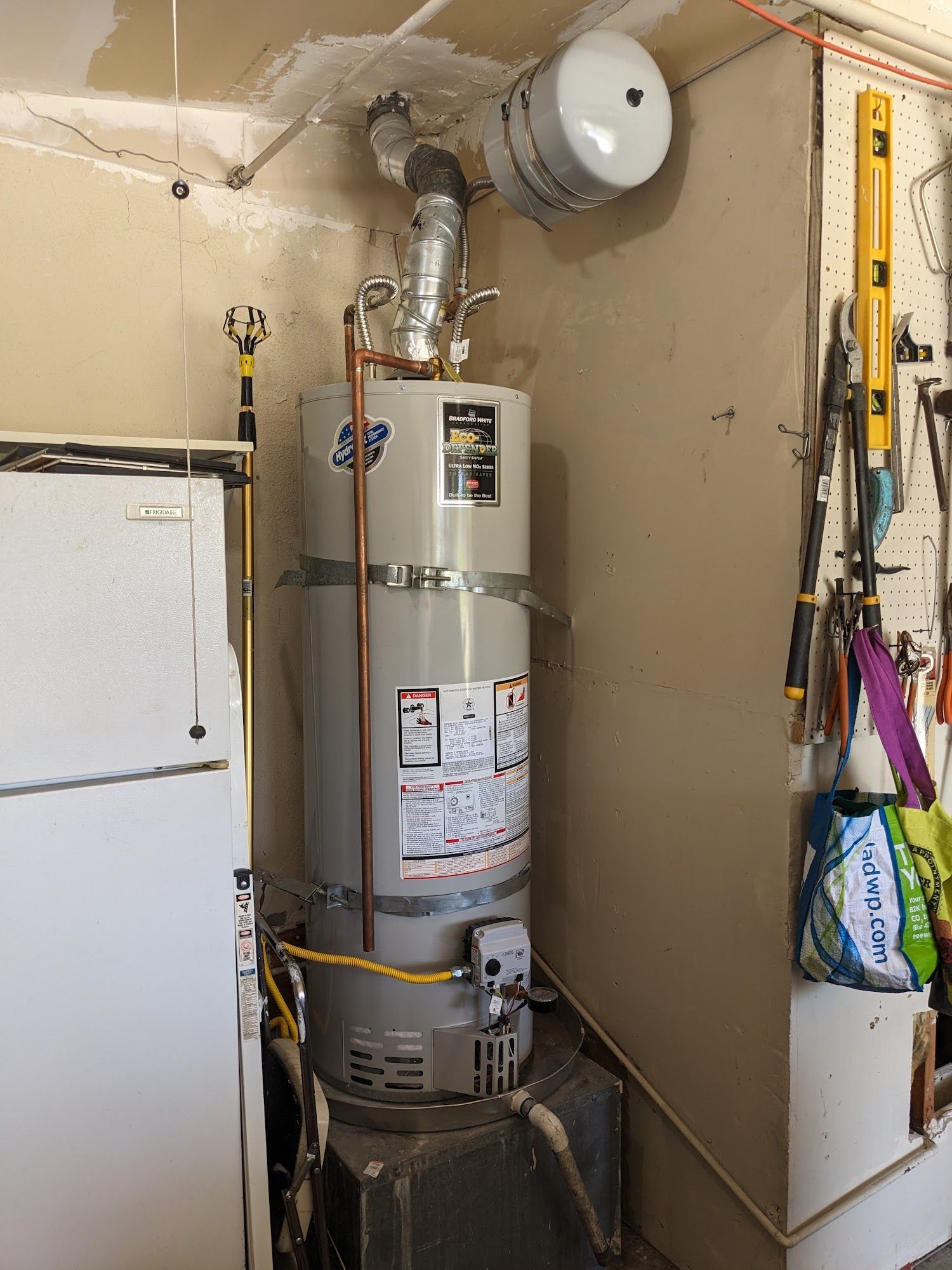 A water heater and attached exhaust system in a corner of a room, next to a refrigerator and wall-mounted tools.