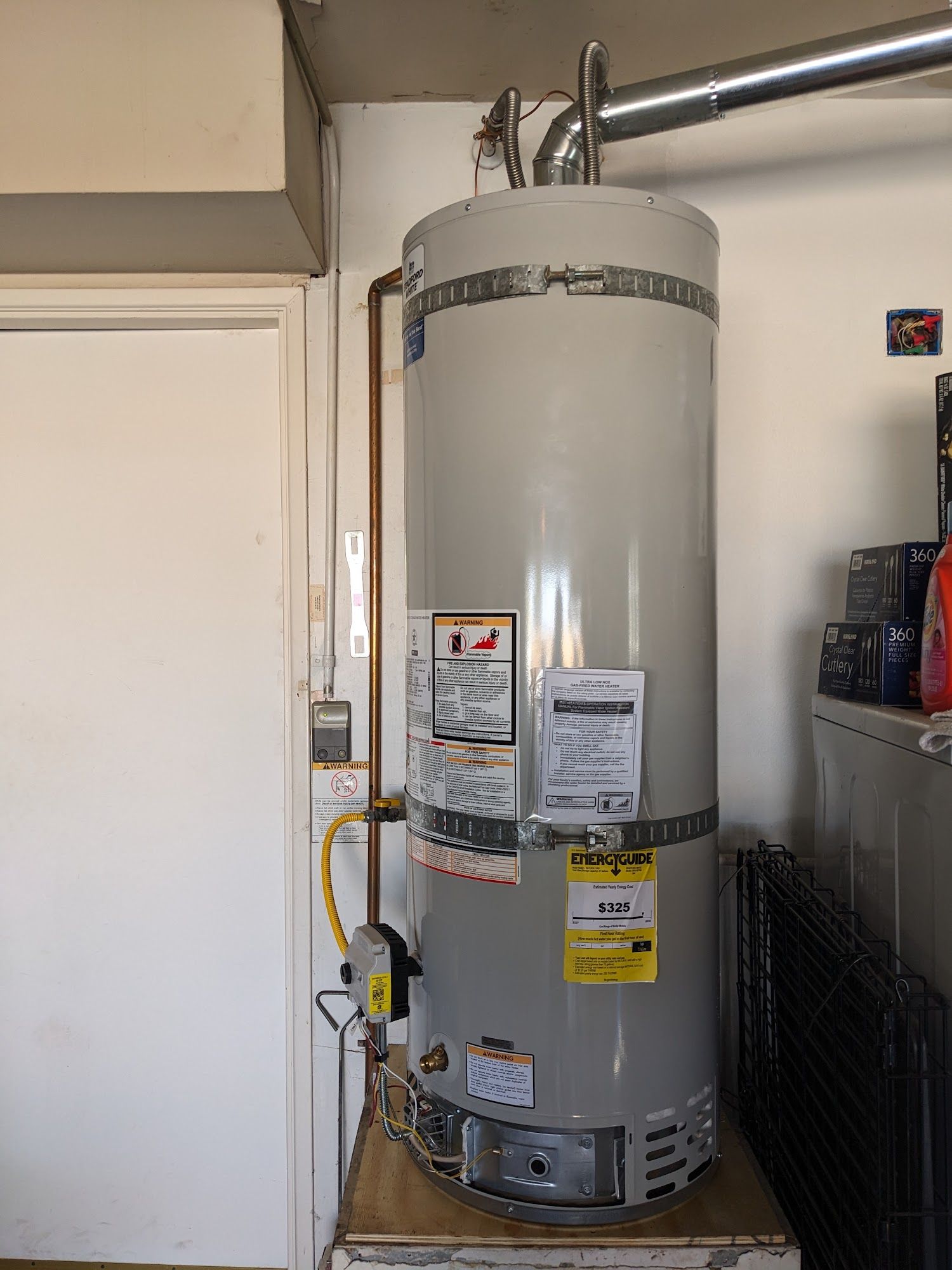 A tall, cylindrical gray water heater in a garage, with copper pipes and a metal vent pipe.