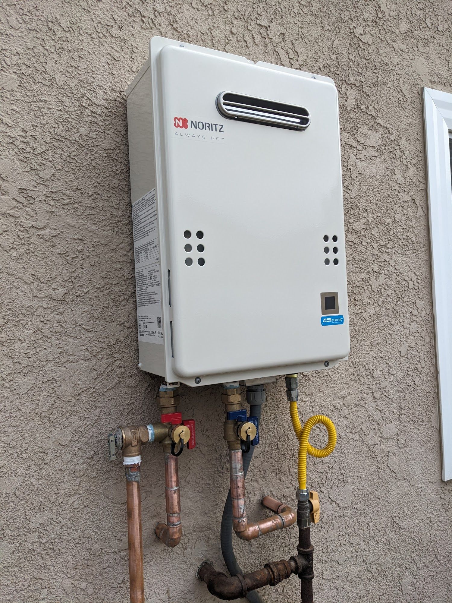 A white tankless water heater mounted on a beige exterior wall with connected pipes.